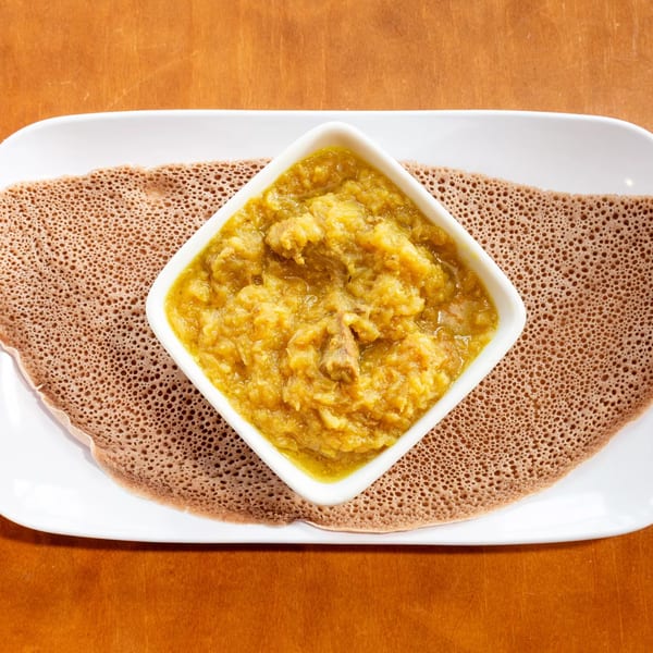 Ethiopian injera bread with a square bowl of yellow stew, possibly shiro wot, on a white plate.