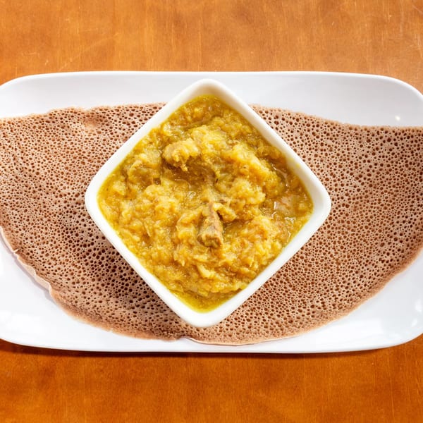 A white square bowl of yellow Ethiopian stew with meat, served on injera bread on a white plate.