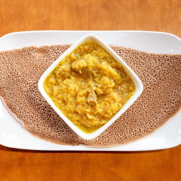 A white square bowl of yellow Ethiopian shiro stew with meat, served on injera bread.